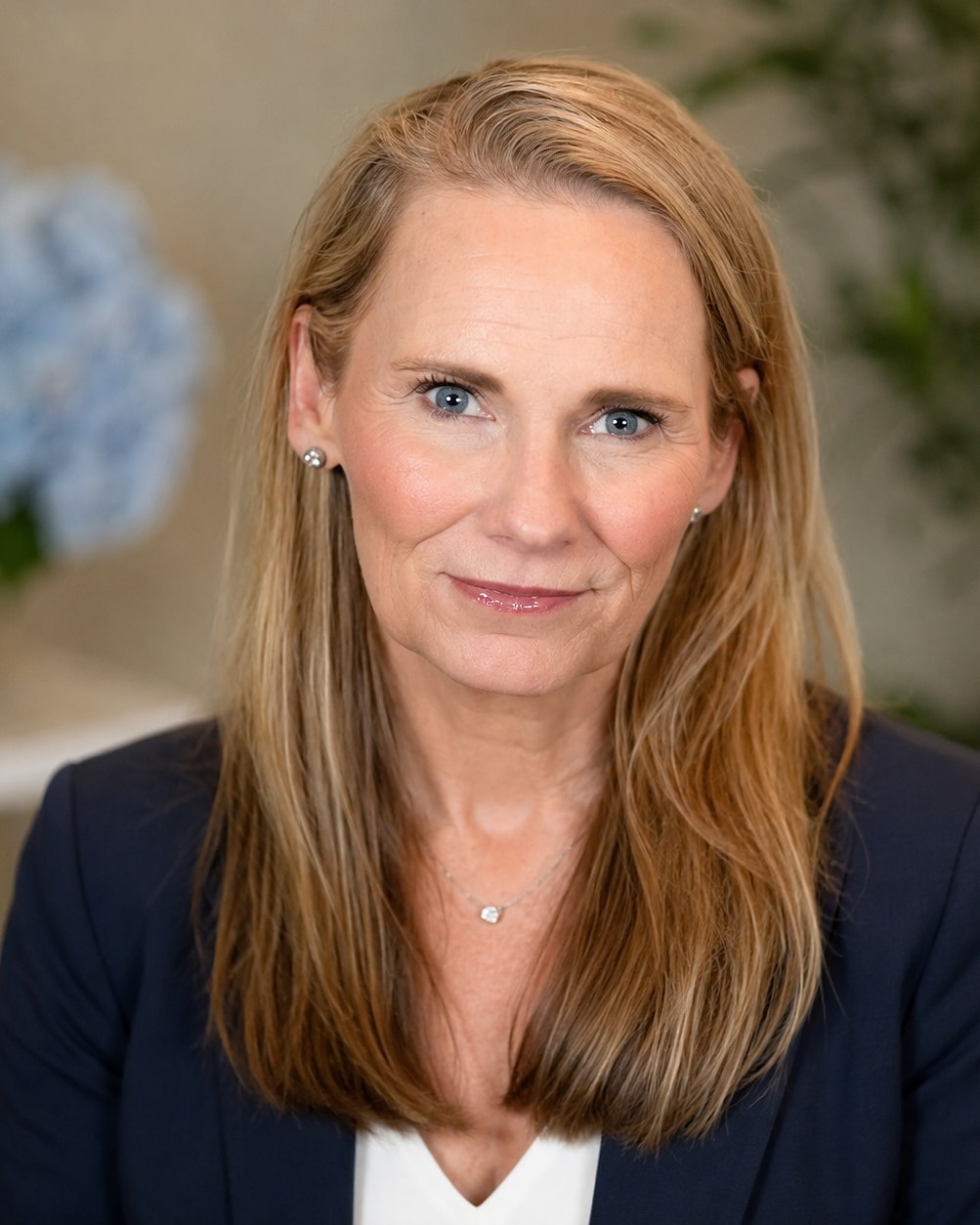 Jennifer Cook, smiling, in a navy blazer, with blue hydrangeas on the table beside her